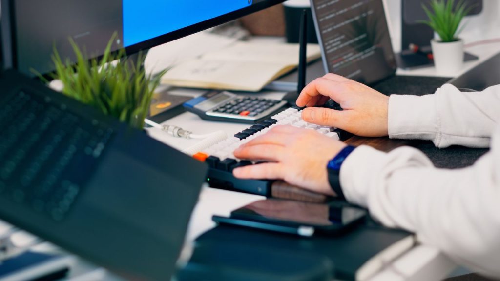 modern-workspace-with-programmer-and-tech-devices-36598848 A close-up of a person typing on a keyboard in a modern tech workspace with gadgets and a monitor.